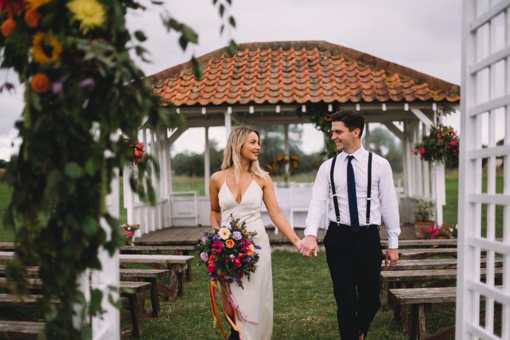 A newly married couple walk away from the Ceremony Pavilion at The Reid Rooms. She has a brightly coloured bouquet and he is wearing black braces. A newly married couple walk away from the Ceremony Pavilion at The Reid Rooms. She has a brightly coloured bouquet and he is wearing black braces.
