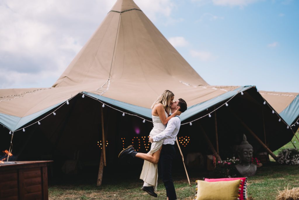 A groom is lifting a bride up to kiss her, standing outside a wedding tipi at The Reid Rooms. A groom is lifting a bride up to kiss her, standing outside a wedding tipi at The Reid Rooms.
