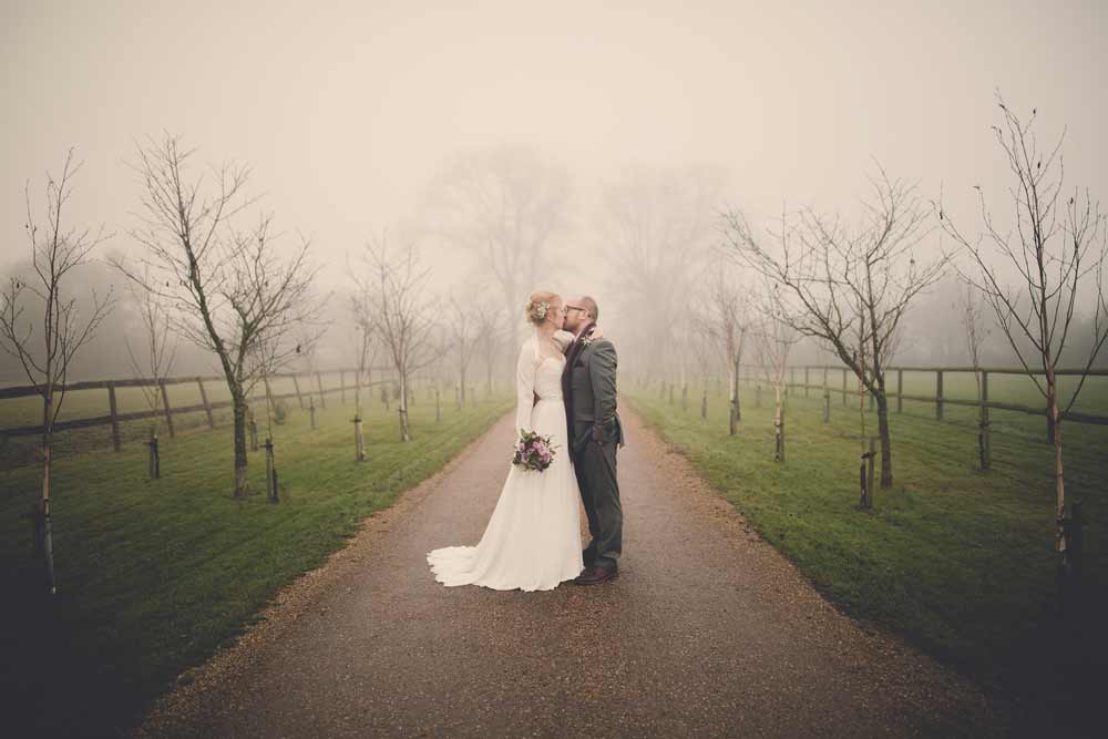 A bride and groom stand and kiss on a misty driveway in the winter at The Reid Rooms. A bride and groom stand and kiss on a misty driveway in the winter at The Reid Rooms.