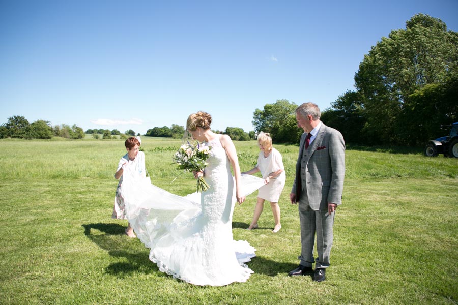 A bride stands in a beautiful sunny green field with her mother, father and a bridesmaid. They are watching her and arranging her train ready for the ceremony at The Reid Rooms. A bride stands in a beautiful sunny green field with her mother, father and a bridesmaid. They are watching her and arranging her train ready for the ceremony at The Reid Rooms.
