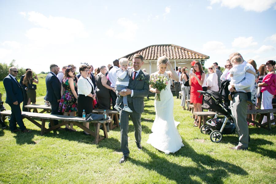 A newly married couple walk through the rows of their wedding guests, walking away from the ceremony pavilion. The sun shines and the fields around them are green and bright. A newly married couple walk through the rows of their wedding guests, walking away from the ceremony pavilion. The sun shines and the fields around them are green and bright.