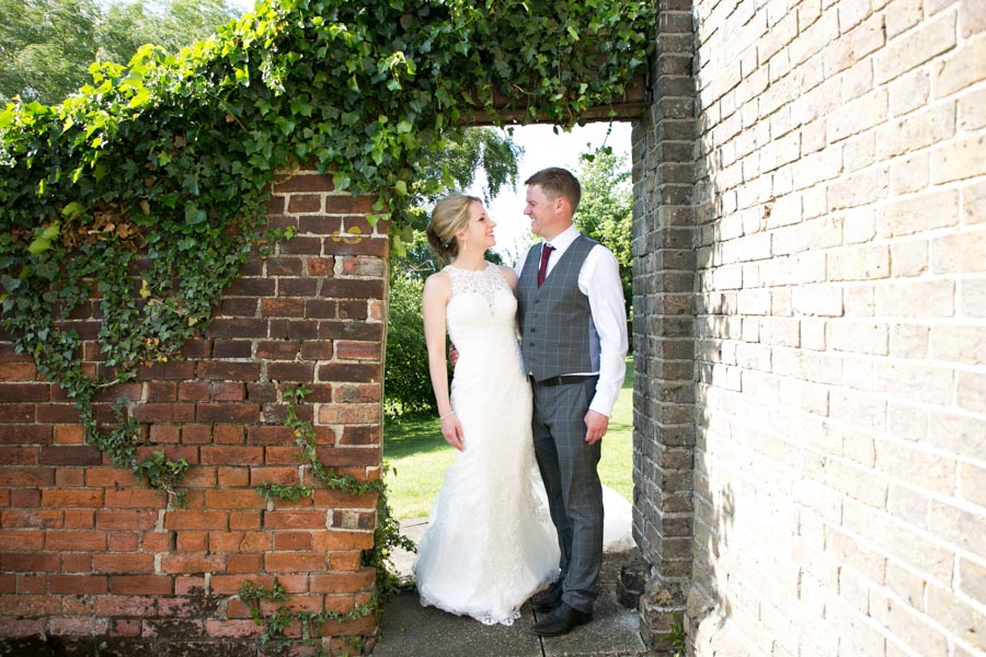 A newly married couple pose in a doorway in a rustic red brick wall covered in trailing green plants. A newly married couple pose in a doorway in a rustic red brick wall covered in trailing green plants.