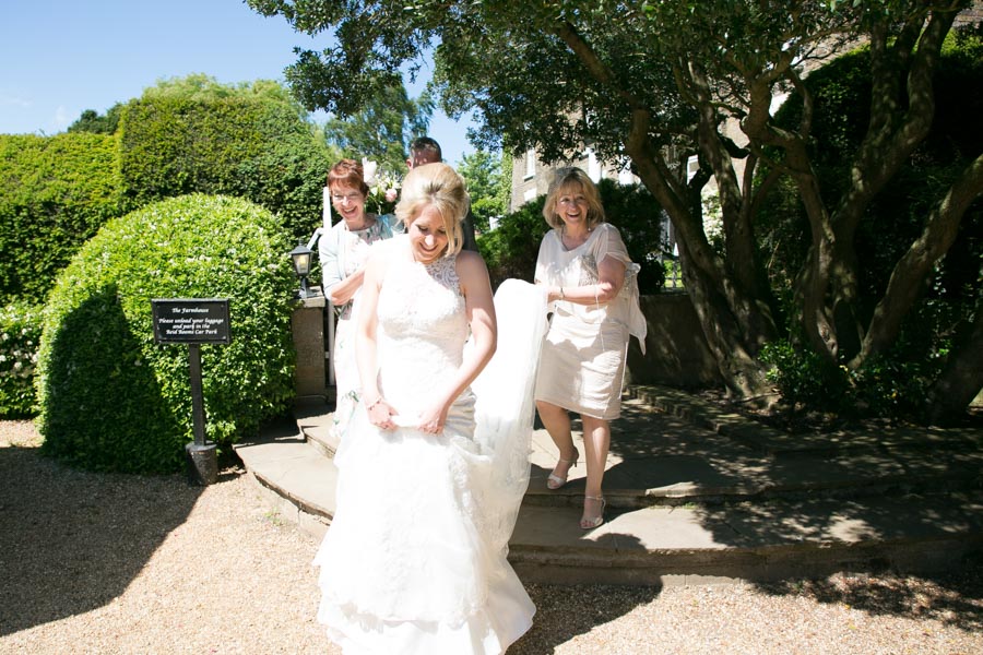Bridal party laughing and leaving Marks Hall Farmhouse on the way to a wedding ceremony at The Reid Rooms. The bride is holding her long train and her mother and bridesmaids are crowded around her. Bridal party laughing and leaving Marks Hall Farmhouse on the way to a wedding ceremony at The Reid Rooms. The bride is holding her long train and her mother and bridesmaids are crowded around her.