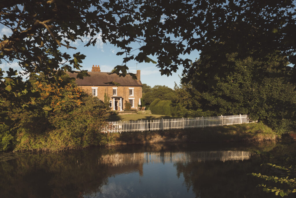 Exterior shot of Marks Hall Farmhouse, the on-site accommodation at The Reid Rooms. A large brick building with a moat and gardens surrounding it.